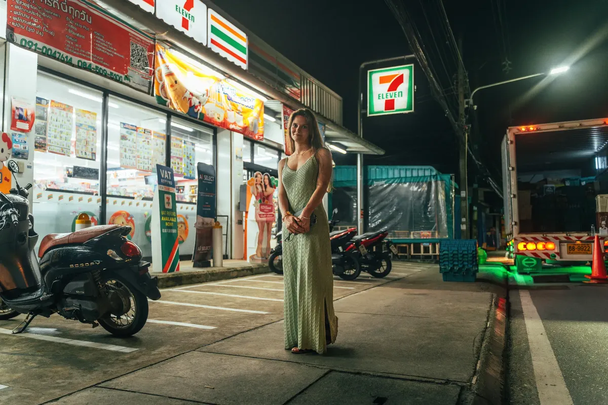 A woman stands outside a 7-Eleven in Phuket at night, surrounded by motorbikes and a parked truck.