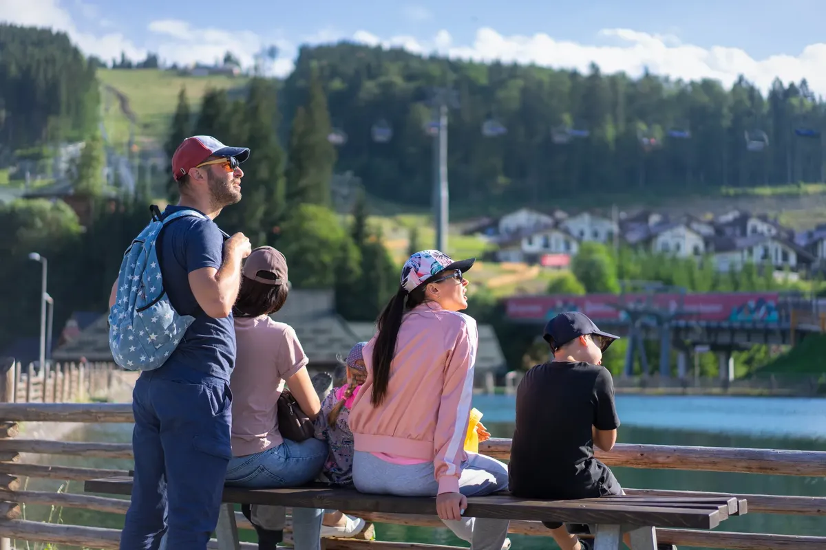 A family enjoys a sunny day sitting on a bench in Bukovel, Ukraine, with scenic mountain views.